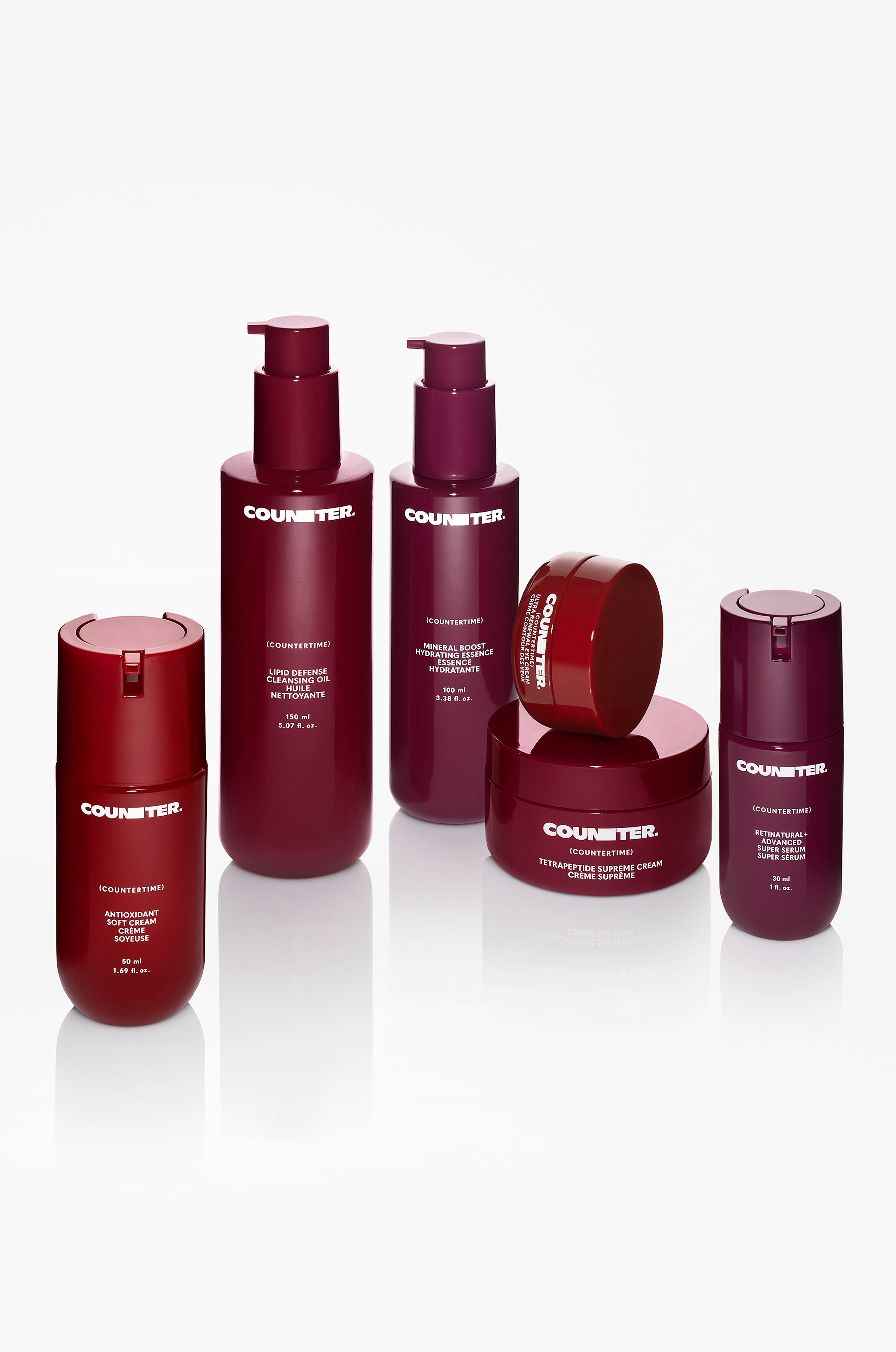 Displays all of the products from the countertime line arranged in a half circle on a mirrored surface. Each bottle is a varying shade of burgundy or bright red.