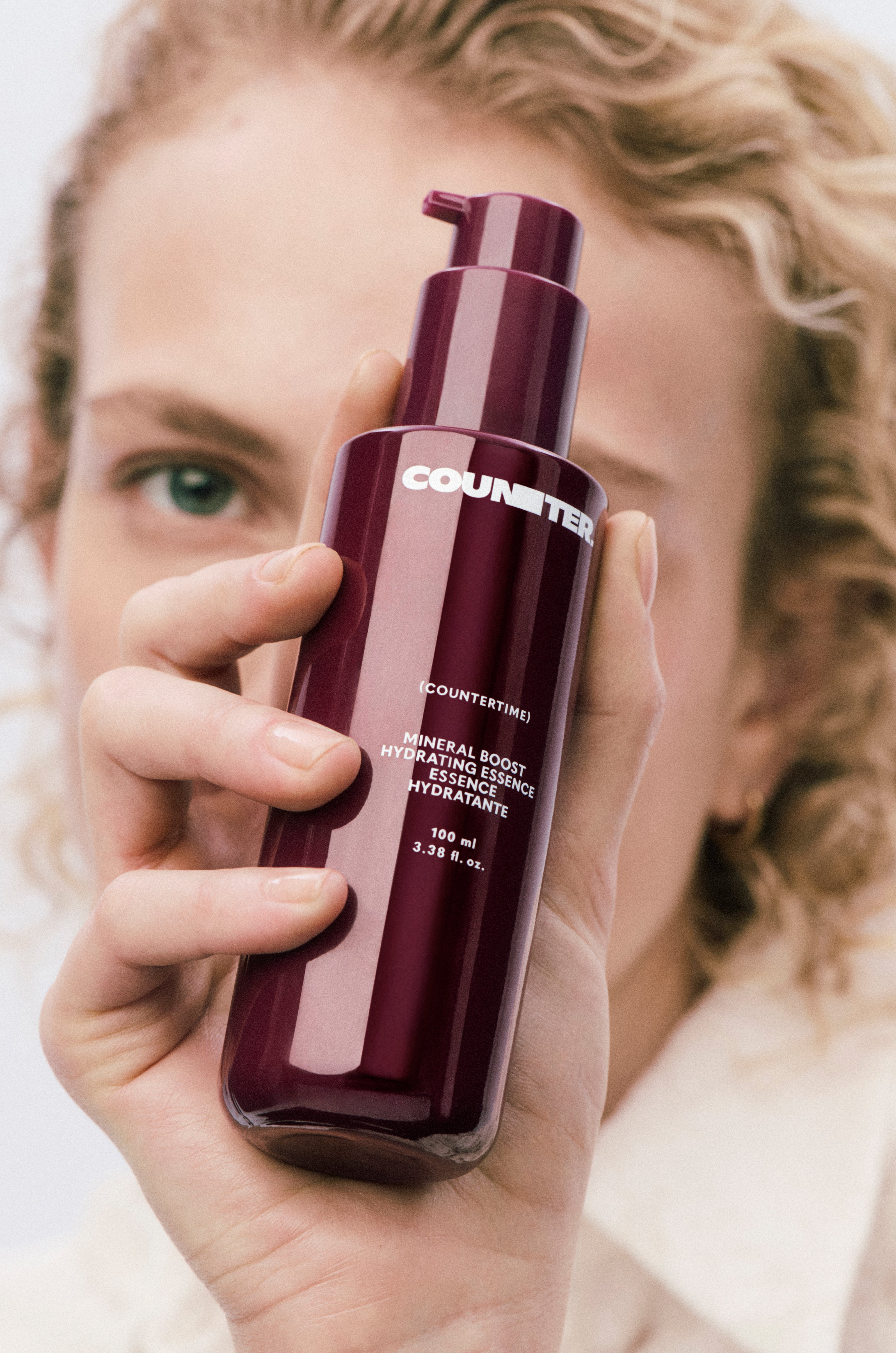Displays a model with light wavy hair holding up a burgundy bottle of countertime mineral boost hydrating essence in front of her face.