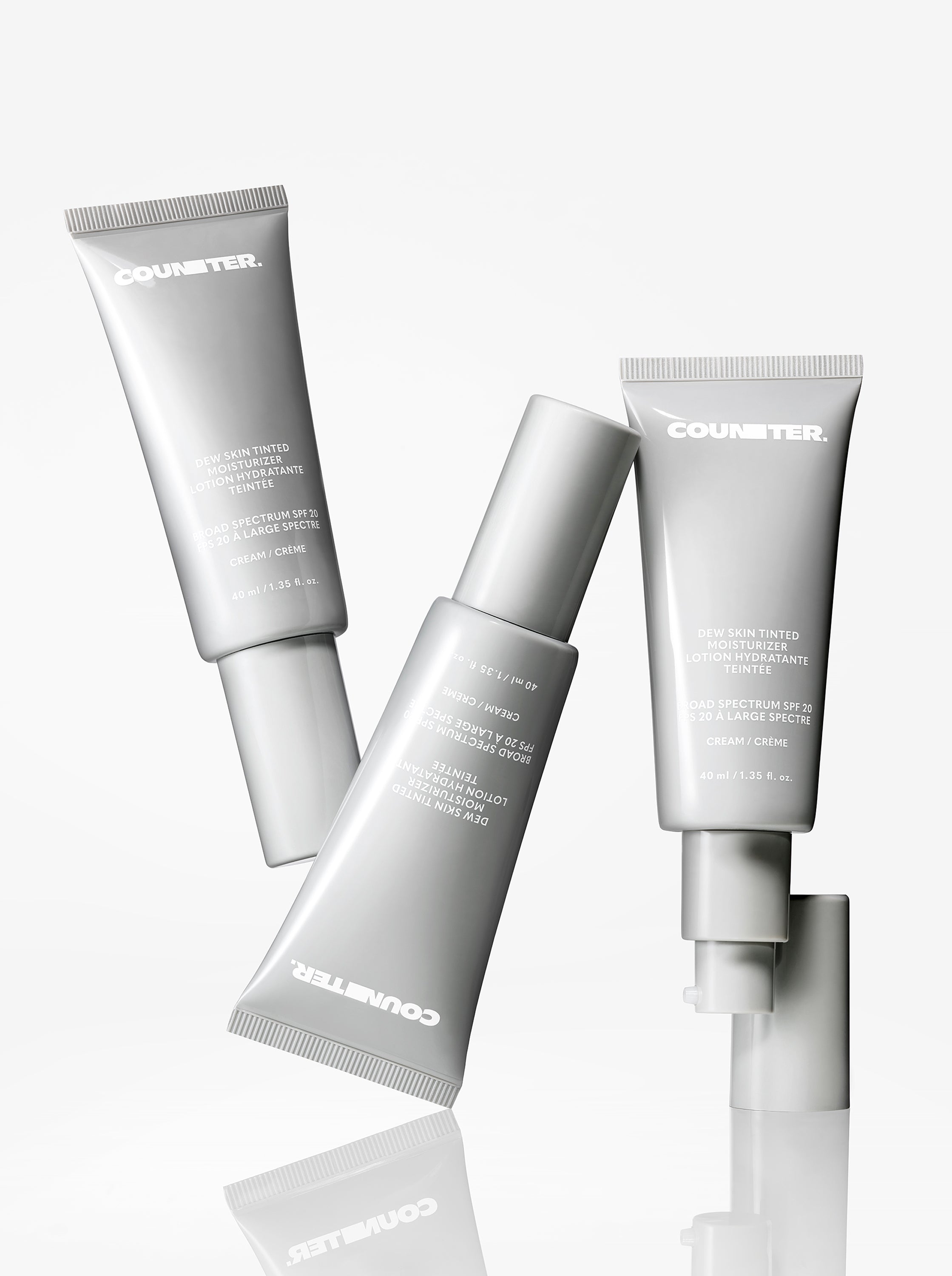 Displays three bottles of the Counter Dew Skin Tinted Moisturizer all tilted in different directions on a mirrored surface.