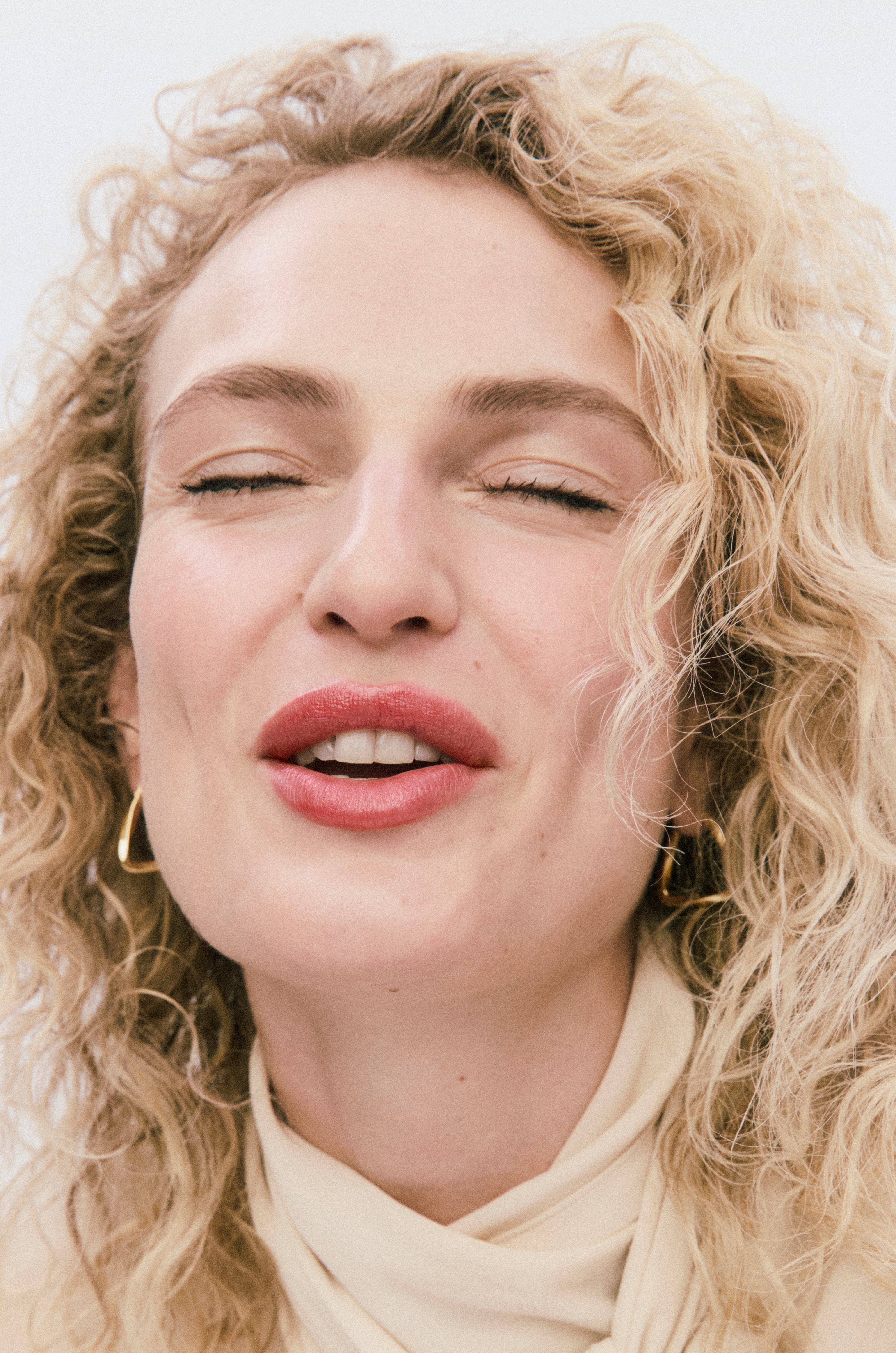 Close-up of a woman with curly blonde hair wearing a beige top.