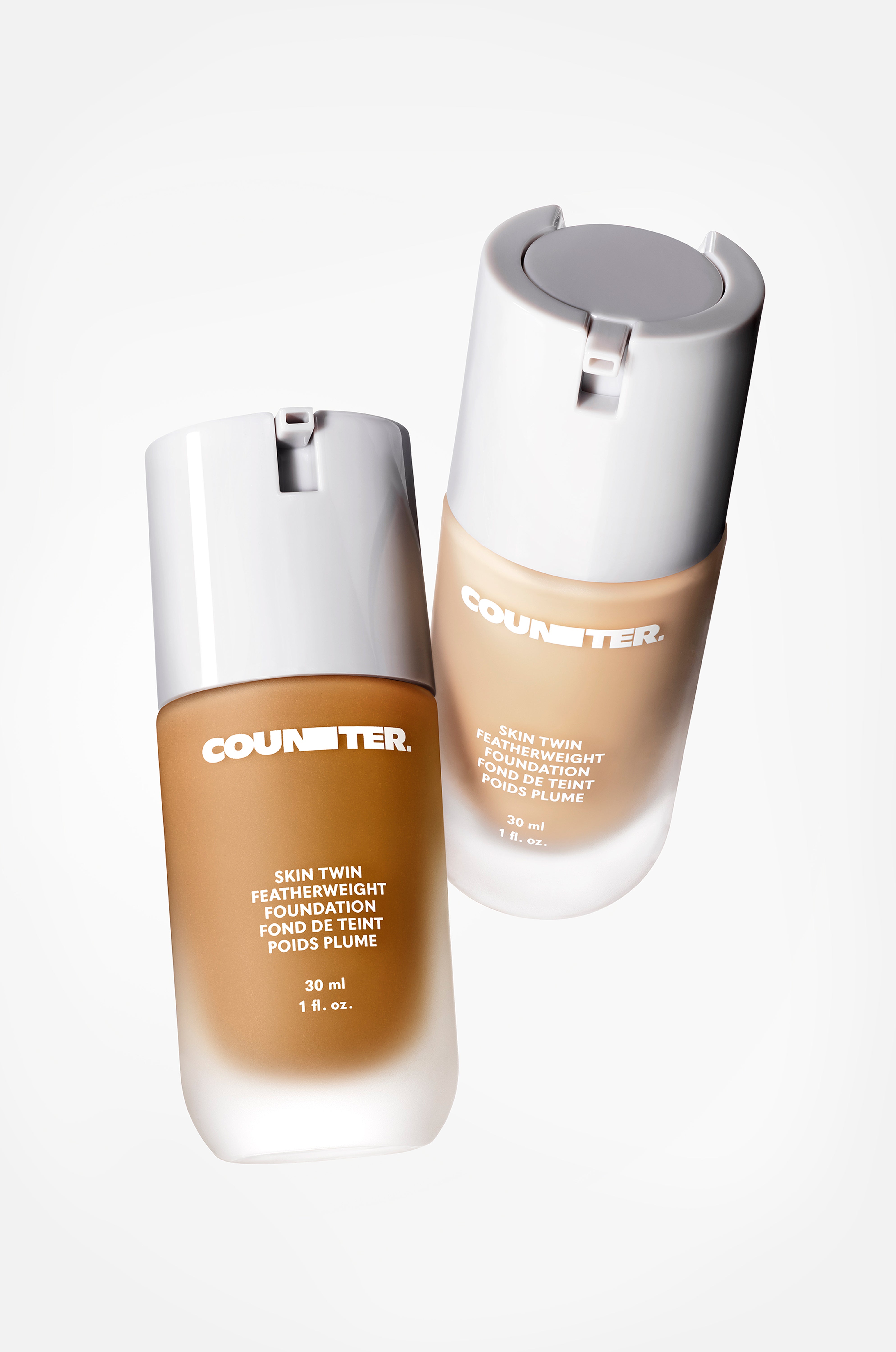 Displays two skin twin featherweight foundations suspended in the air in front of a white background. The componentry is glass with a white lid. There is one darker and one lighter shade featured.
