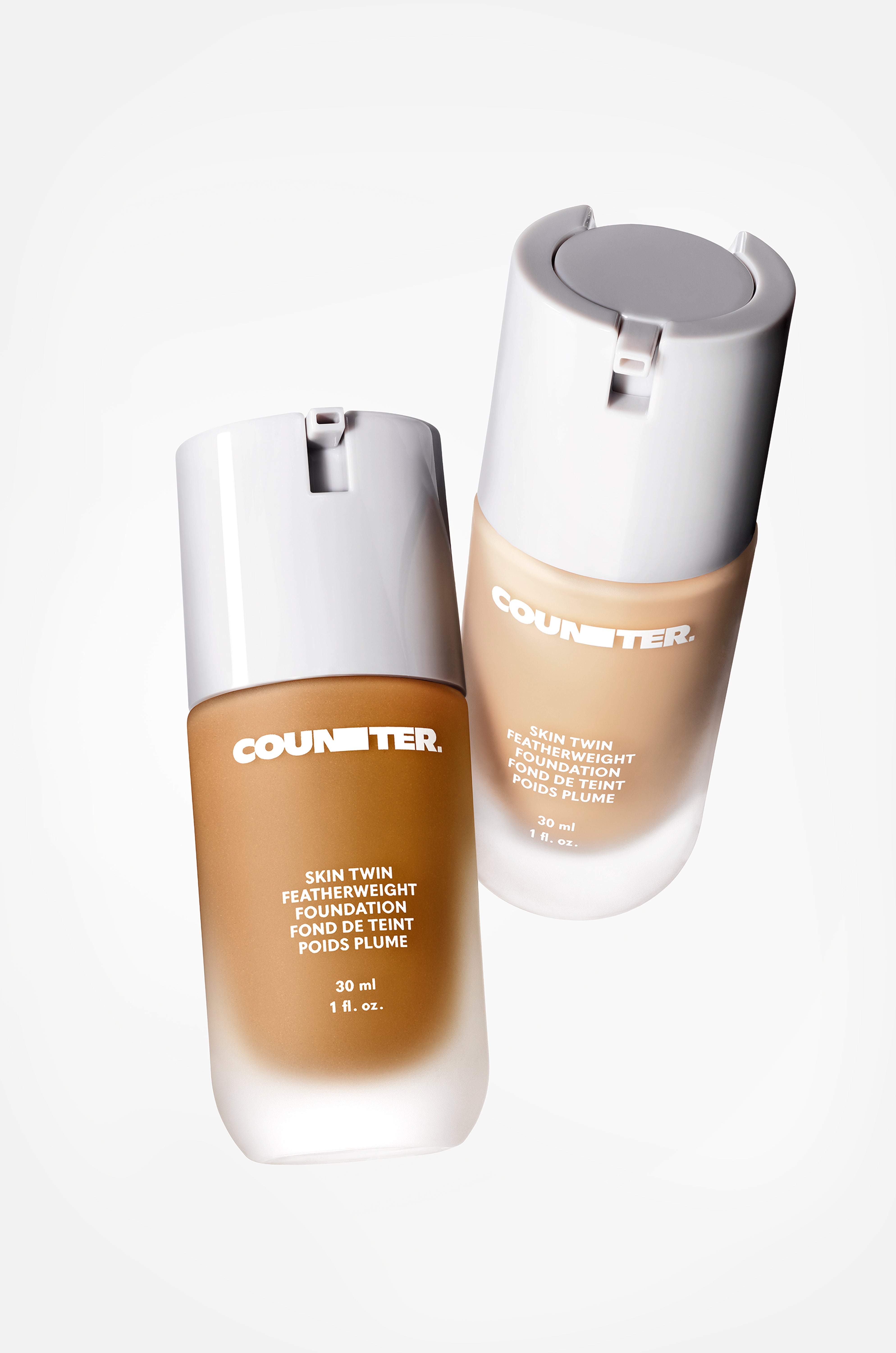 Displays two skin twin featherweight foundations suspended in the air in front of a white background. The componentry is glass with a white lid. There is one darker and one lighter shade featured.
