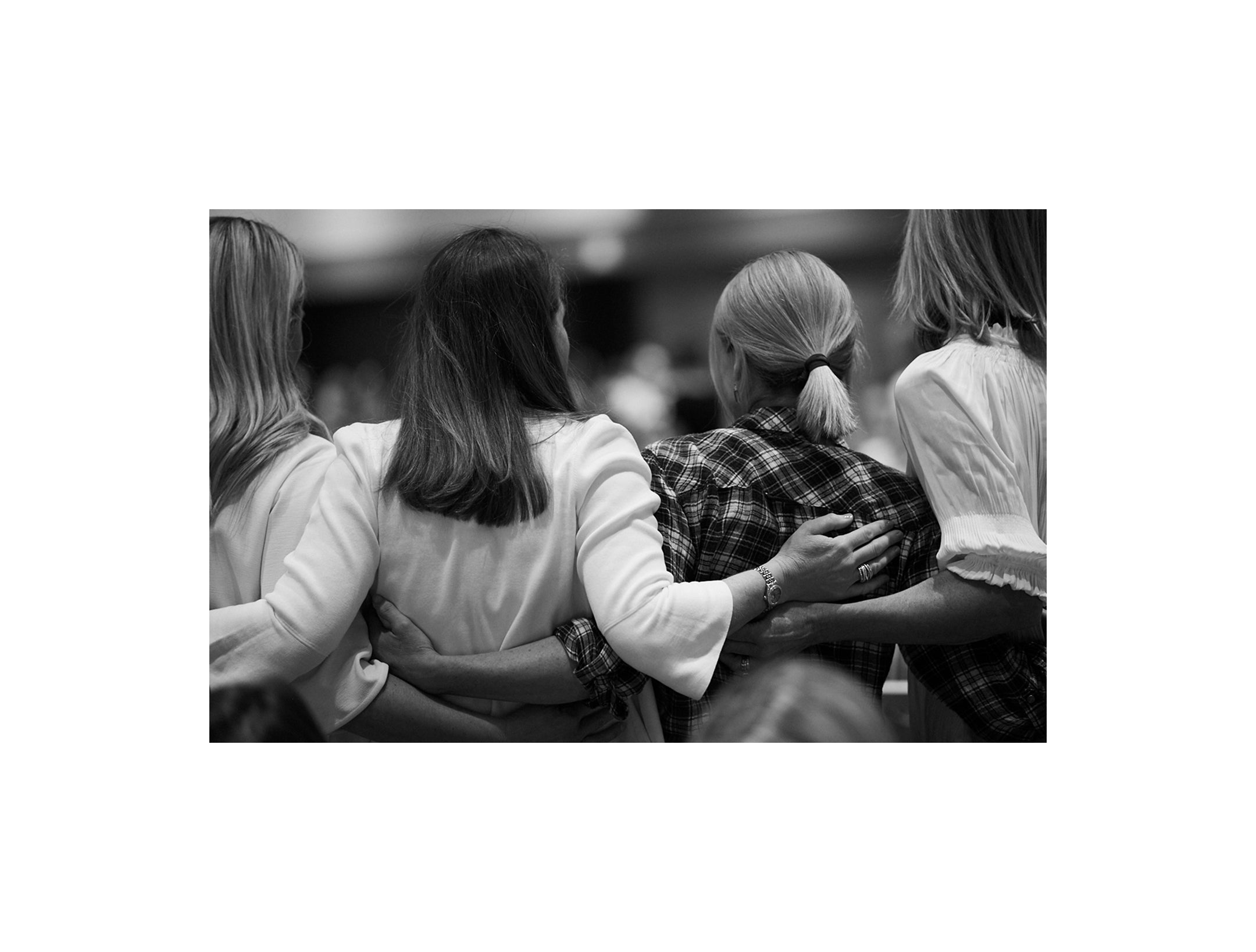 Displays a group of women from behind with their arms around one another.