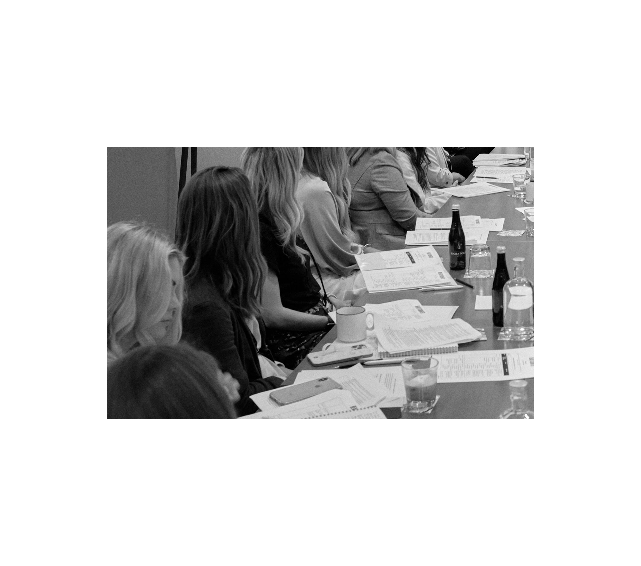 Displays a group of women looking at documents on a table in front of them.