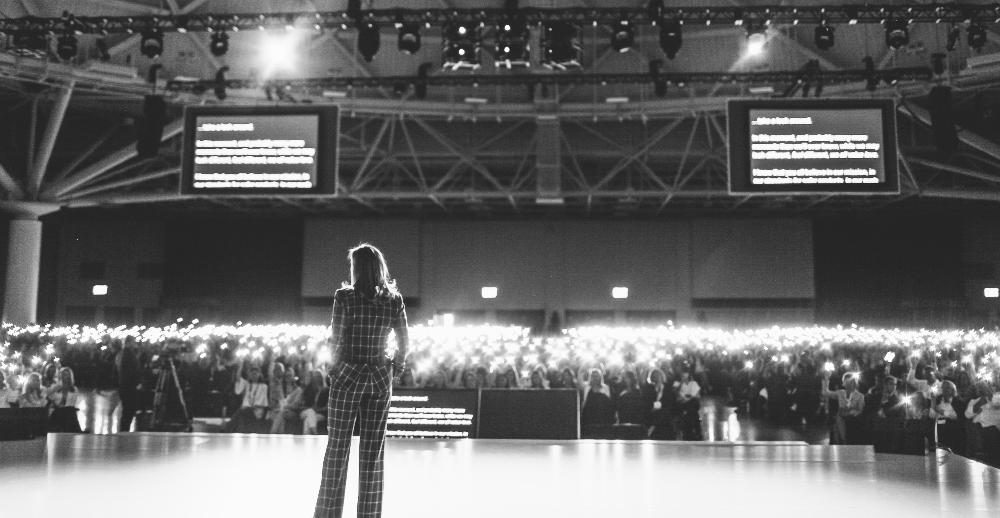 Displays Gregg Renfrew wearing a checkered suit on stage speaking into a microphone. She is facing a crowd filled with people whose phones are illuminated like lights.