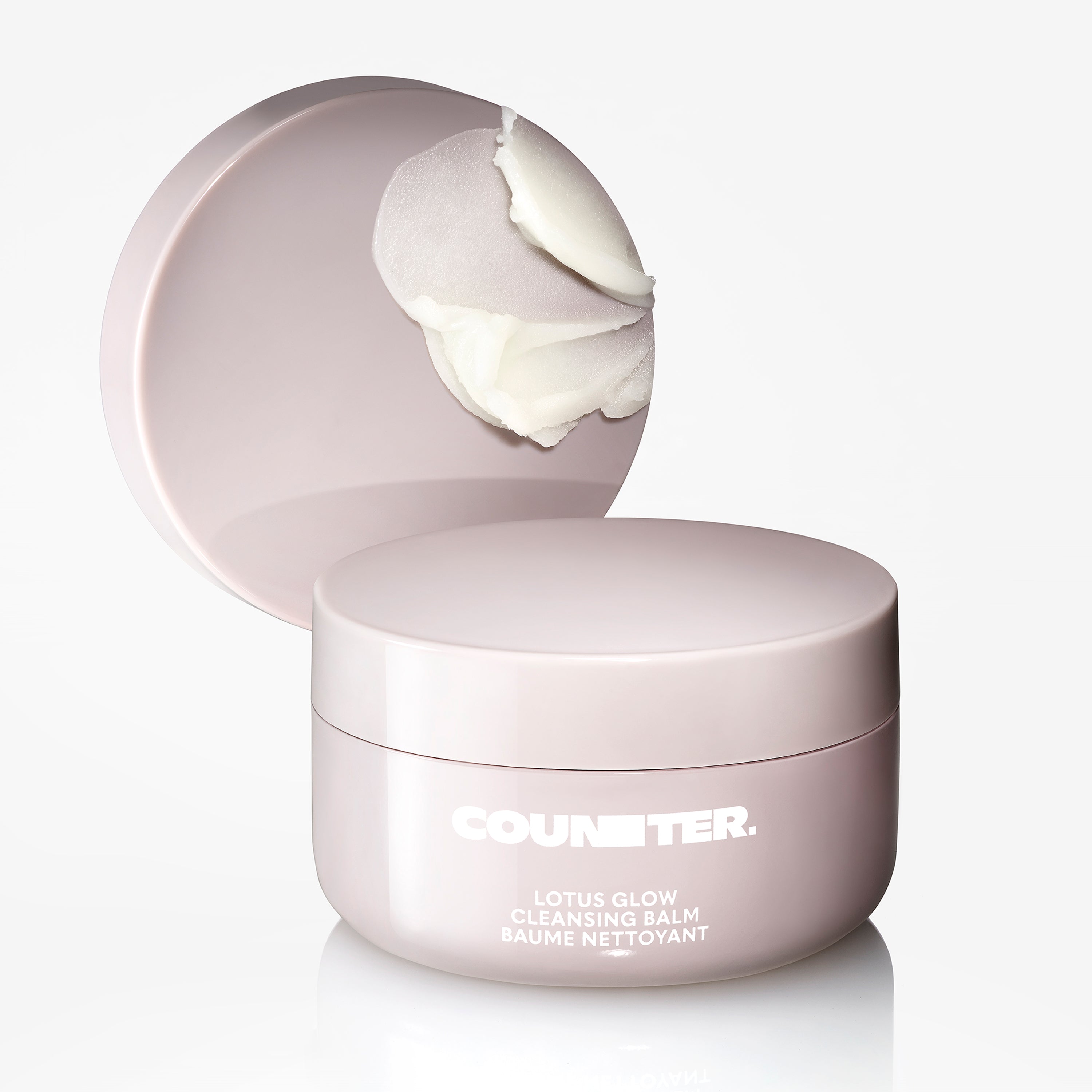 Displays a Counter Lotus Glow Cleansing Balm in a pale lavender jar with the product smear on a lid displayed behind the jar. 