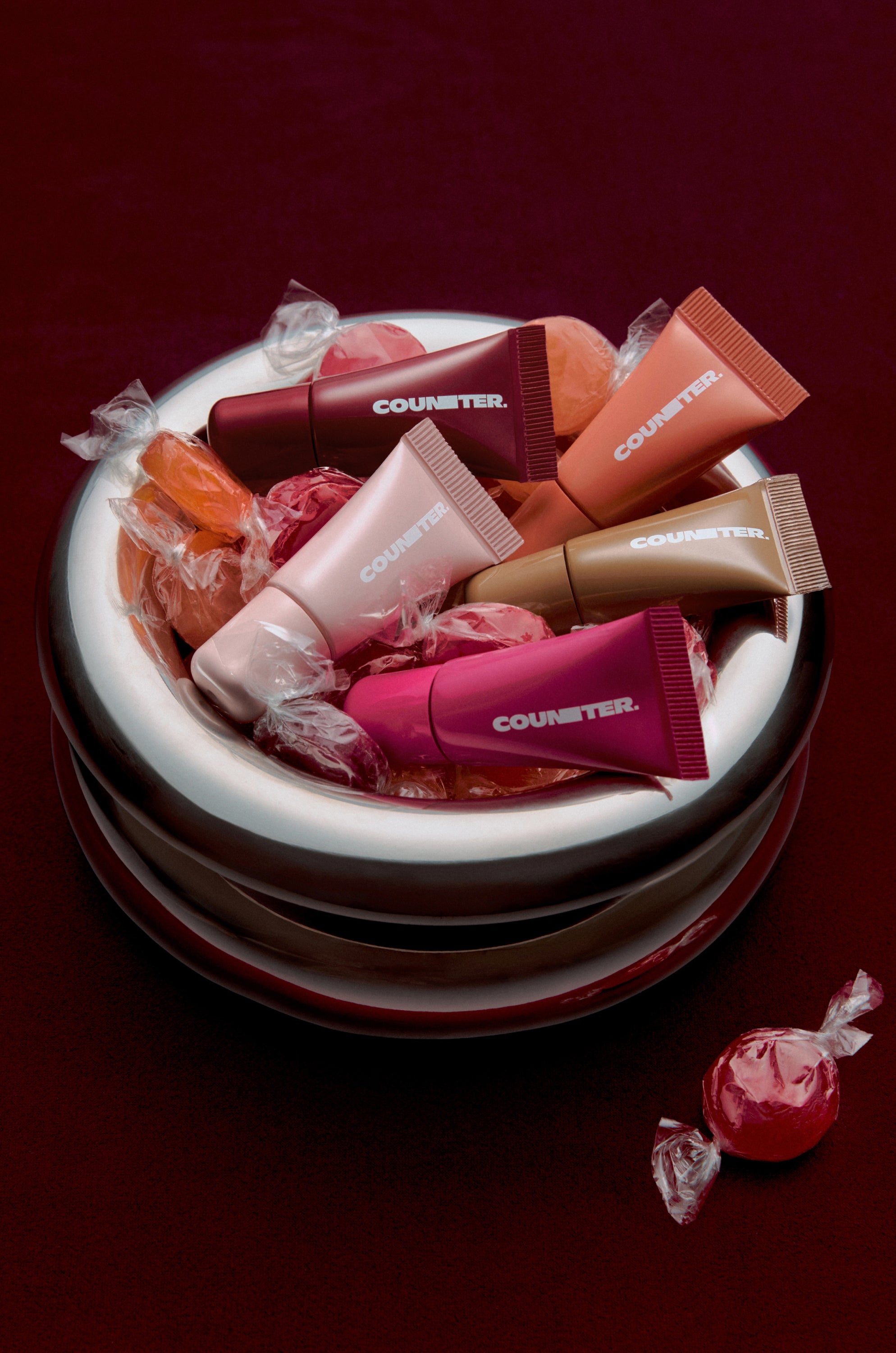 A styled product shot of several Counter lip balm tubes in assorted shades, arranged inside a white bowl filled with wrapped red and pink candies on a dark burgundy background.